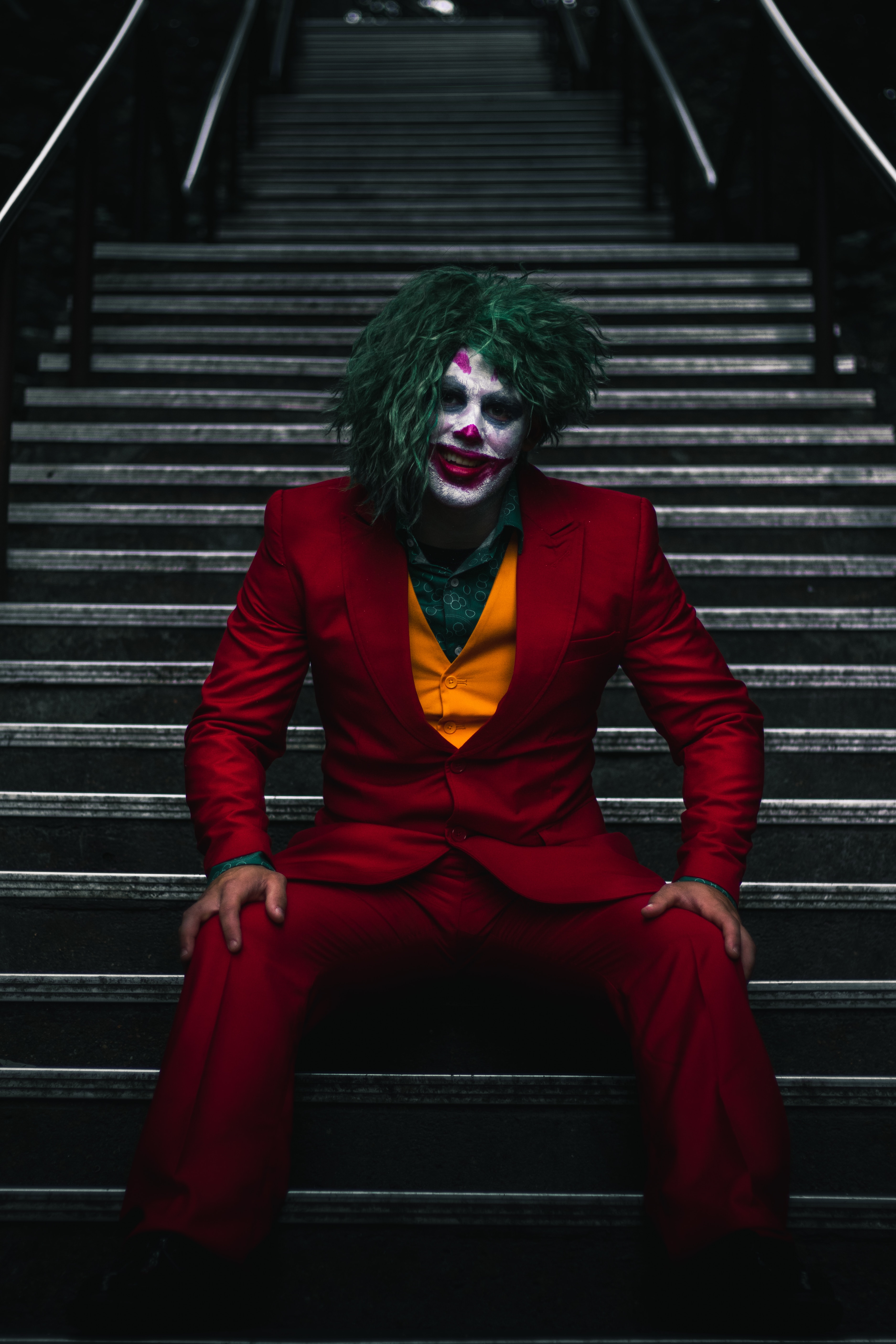 A clown with green hair sitting on stairs.